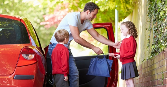 dad with two kids standing outside of an open car door, both kids are wearing school uniforms and he is handing a backpack to the little girl