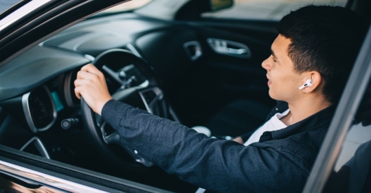 man sitting in the driver's seat of a small car with his left hand on the wheel, he is wearing a t-shirt and open button down top and has Apple AirPods in his ear